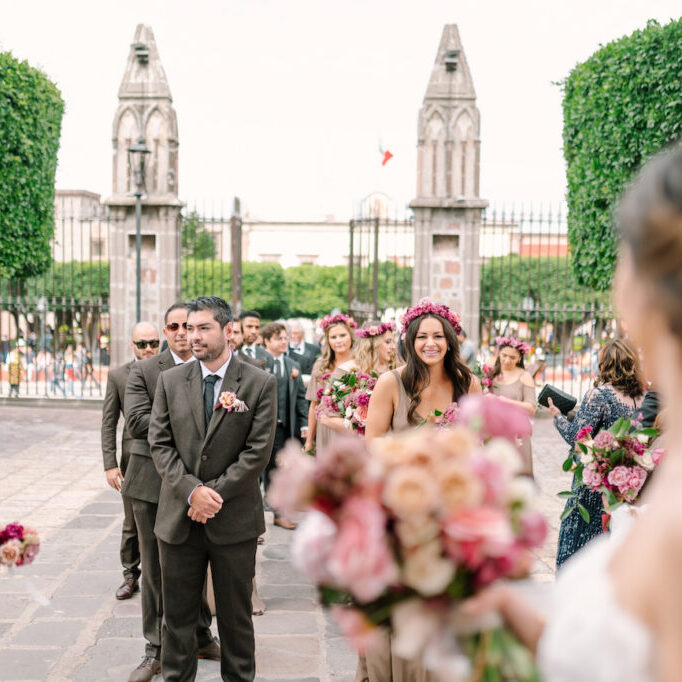 bridesmaids with flower crowns and groomsmen in grey suits look at bride and groom entering San Miguel chapel