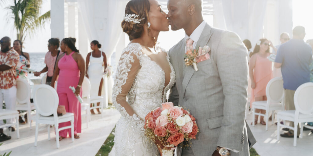 A bride and groom kiss at their wedding ceremony, with the bride holding a bouquet of pink roses and guests seated in the background.