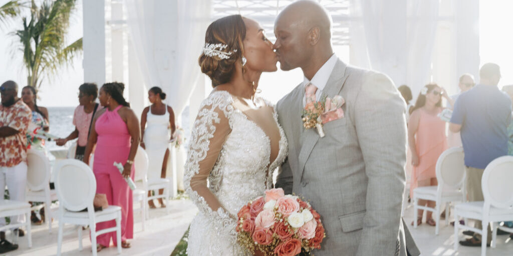 A bride and groom kiss at their wedding ceremony, with the bride holding a bouquet of pink roses and guests seated in the background.