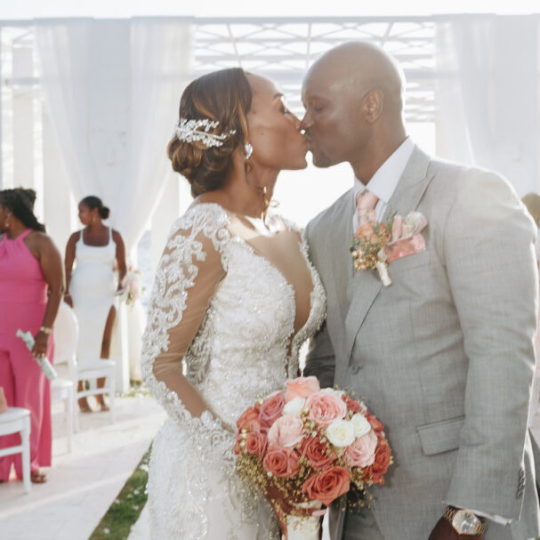 A bride and groom kiss at their wedding ceremony, with the bride holding a bouquet of pink roses and guests seated in the background.